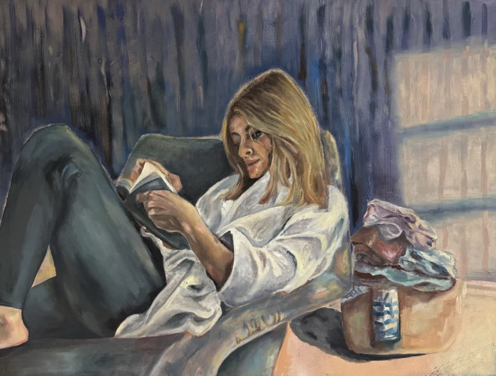 A woman in a white robe curls up in an armchair reading a book, with a laundry basket beside her in soft blue light.